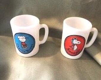 Può includere: Due tazze in vetro opalino bianco con personaggi Peanuts. La tazza a sinistra presenta un grafico circolare blu di Charlie Brown con il testo Charlie Brown. La tazza a destra ha un grafico circolare rosso di Snoopy con il testo Snoopy.