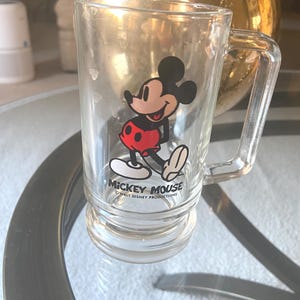 May include: Clear glass mug featuring Mickey Mouse. The cartoon character is depicted in his classic outfit with red shorts, white gloves, and yellow shoes. The mug has a handle and the text "Mickey Mouse" and "Walt Disney Productions" printed on it.