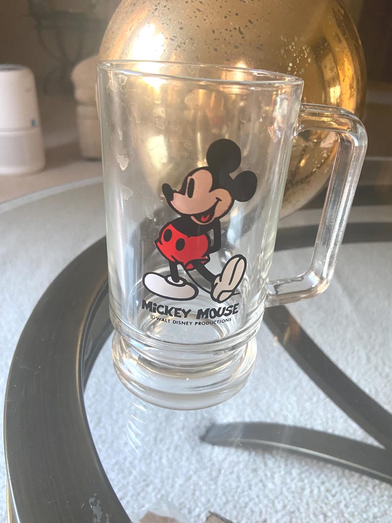 May include: Clear glass mug featuring Mickey Mouse. The cartoon character is depicted in his classic outfit with red shorts, white gloves, and yellow shoes. The mug has a handle and the text "Mickey Mouse" and "Walt Disney Productions" printed on it.