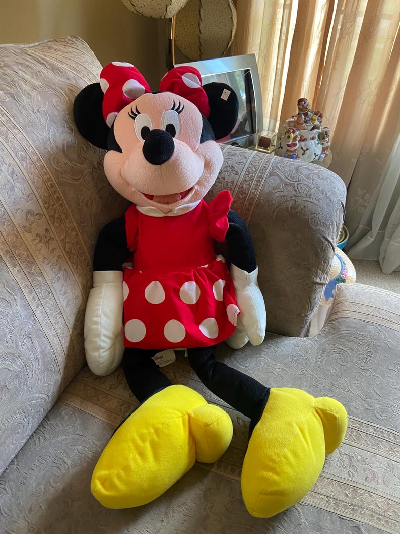 May include: A large Minnie Mouse plush doll. The doll has a red dress with white polka dots, a matching bow, and yellow shoes. The doll's face is pink with black eyes, a black nose, and black ears. The doll is sitting on a patterned couch.