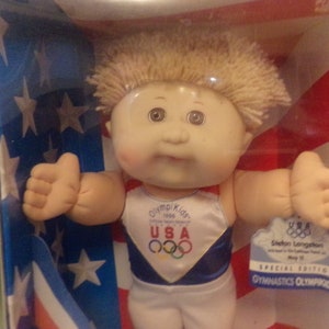 May include: A Cabbage Patch Kid doll dressed in a blue and white gymnastics leotard with the words "Olympic Kids 1996 Official Team Mascot" and "USA" printed on the front. The doll is holding a small tag that reads "USA 1996 Sheldon Langston with a 10-day qualifying vault on May 10 Special Edition Gymnastics Olympics". The doll is posed in front of an American flag.