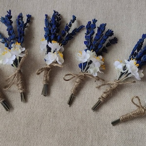Peut inclure: Six petits bouquets de lavande séchée et de fleurs blanches liés avec de la ficelle. Les bouquets sont disposés en rangée sur un fond de tissu beige.