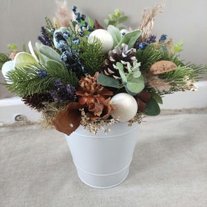 Könnte beinhalten: Ein dekoratives Arrangement aus künstlichem Grün, Tannenzapfen, Beeren und weißen Kugeln in einem weißen Metalleimer. Das Arrangement umfasst Lavendelzweige, salbeigrüne Blätter und braune Akzente.