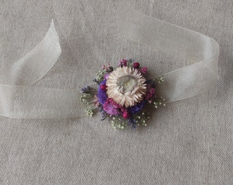 May include: A small bouquet of dried flowers in shades of pink, purple, and white, tied with a sheer white ribbon. The flowers are arranged in a circular shape and are accented with small green leaves.