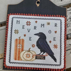 May include: A cross-stitch wall hanging featuring a black raven perched on a sign labeled 1845, next to two pumpkins. The design includes the text NEVERMORE, small star motifs, and acorns, all bordered by a black frame with orange pom-pom trim.