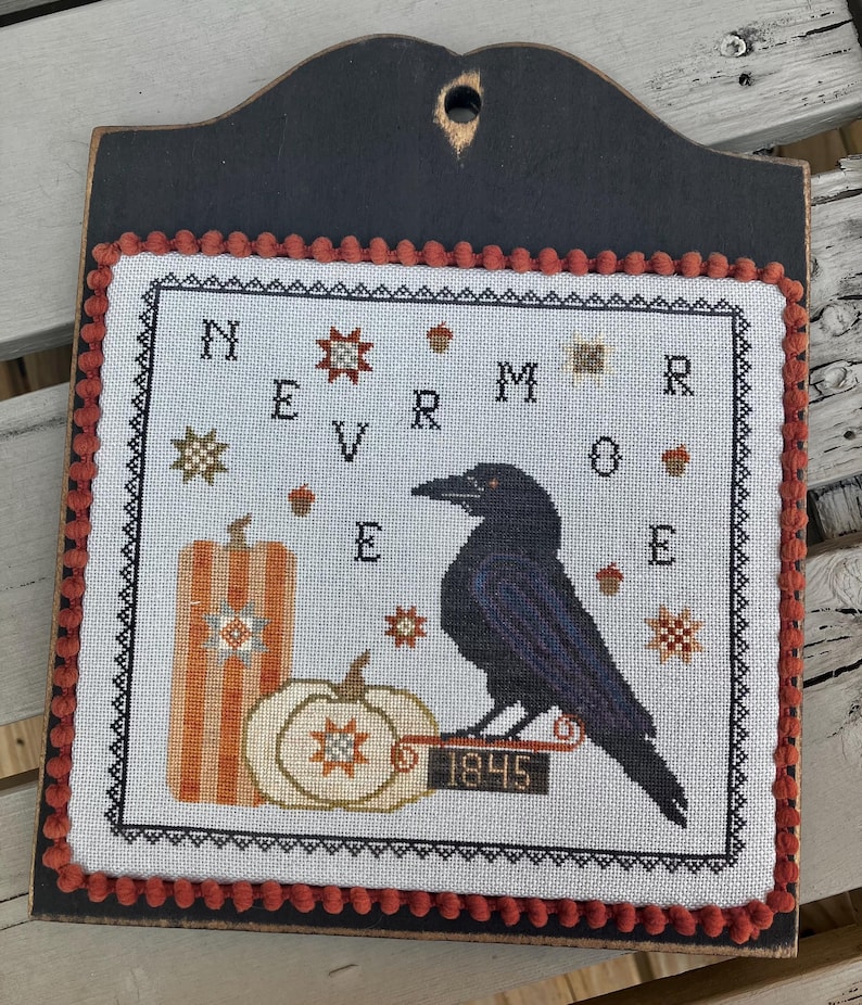 May include: A cross-stitch wall hanging featuring a black raven perched on a sign labeled 1845, next to two pumpkins. The design includes the text NEVERMORE, small star motifs, and acorns, all bordered by a black frame with orange pom-pom trim.
