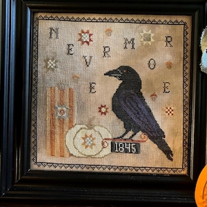 May include: A framed cross-stitch embroidery featuring a black raven perched on a sign labeled 1845, next to a white pumpkin and a striped pillar. The text NEVERMORE is stitched in black letters, surrounded by small star and acorn motifs on beige fabric.