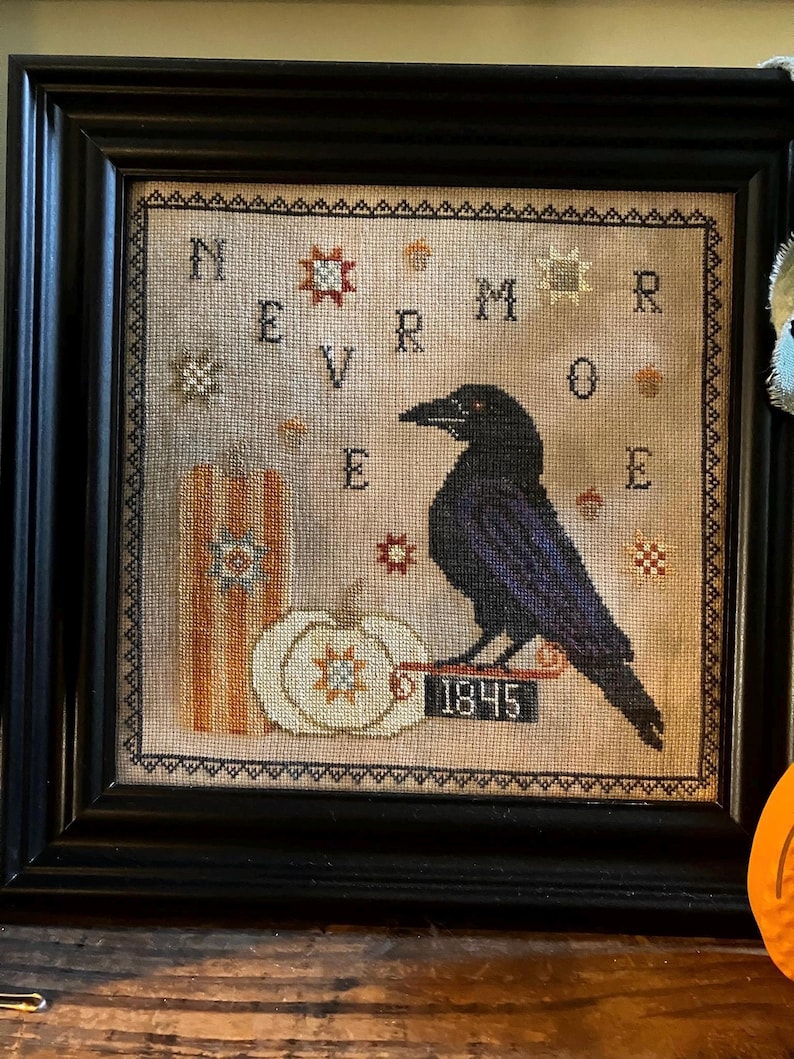 May include: A framed cross-stitch embroidery featuring a black raven perched on a sign labeled 1845, next to a white pumpkin and a striped pillar. The text NEVERMORE is stitched in black letters, surrounded by small star and acorn motifs on beige fabric.