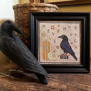 May include: A framed cross-stitch sampler featuring a black raven, a pumpkin, and the text NEVERMORE 1845, displayed alongside a black fabric raven figurine. The sampler uses a neutral fabric background with orange, black, and white thread accents.