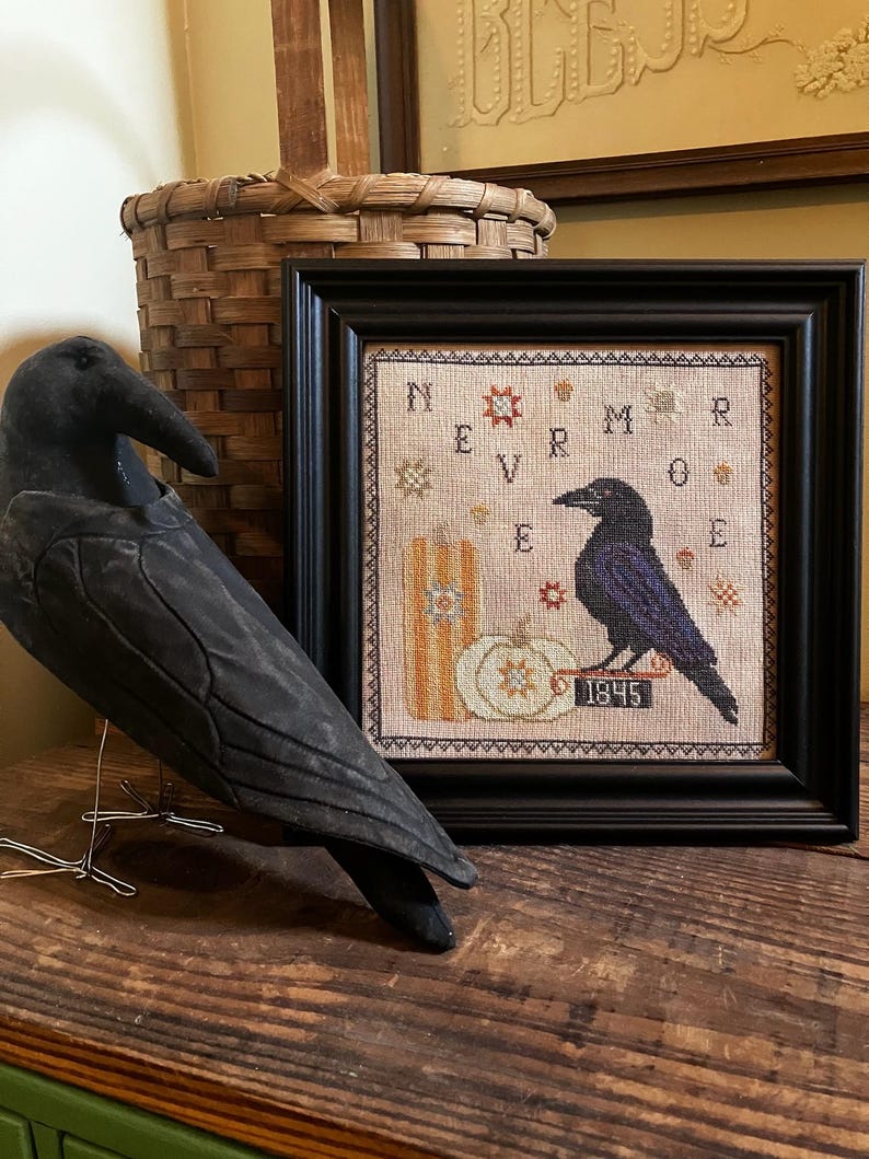 May include: A framed cross-stitch sampler featuring a black raven, a pumpkin, and the text NEVERMORE 1845, displayed alongside a black fabric raven figurine. The sampler uses a neutral fabric background with orange, black, and white thread accents.