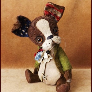 May include: A handmade plush toy dog with brown and white fur, wearing a green jacket and a red, white, and blue fabric bow tie. The dog has a key attached to its neck and is sitting on a brown surface. The toy is labeled "Alla Bears".