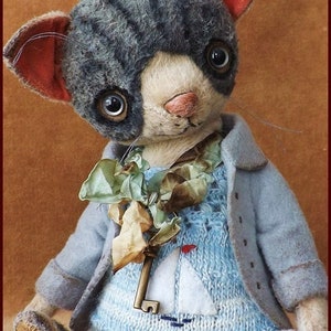 May include: A gray and white stuffed cat wearing a blue knitted sweater with a sailboat design and a brown and green ribbon around its neck. The cat is holding a gold key in its paw.