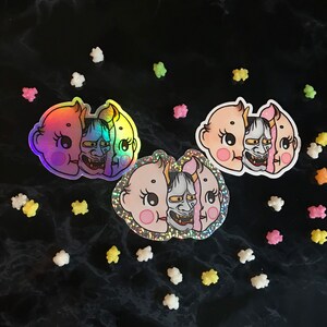 May include: Three iridescent stickers featuring a baby face with a demon mask. The stickers are on a black background with white and pink sprinkles.