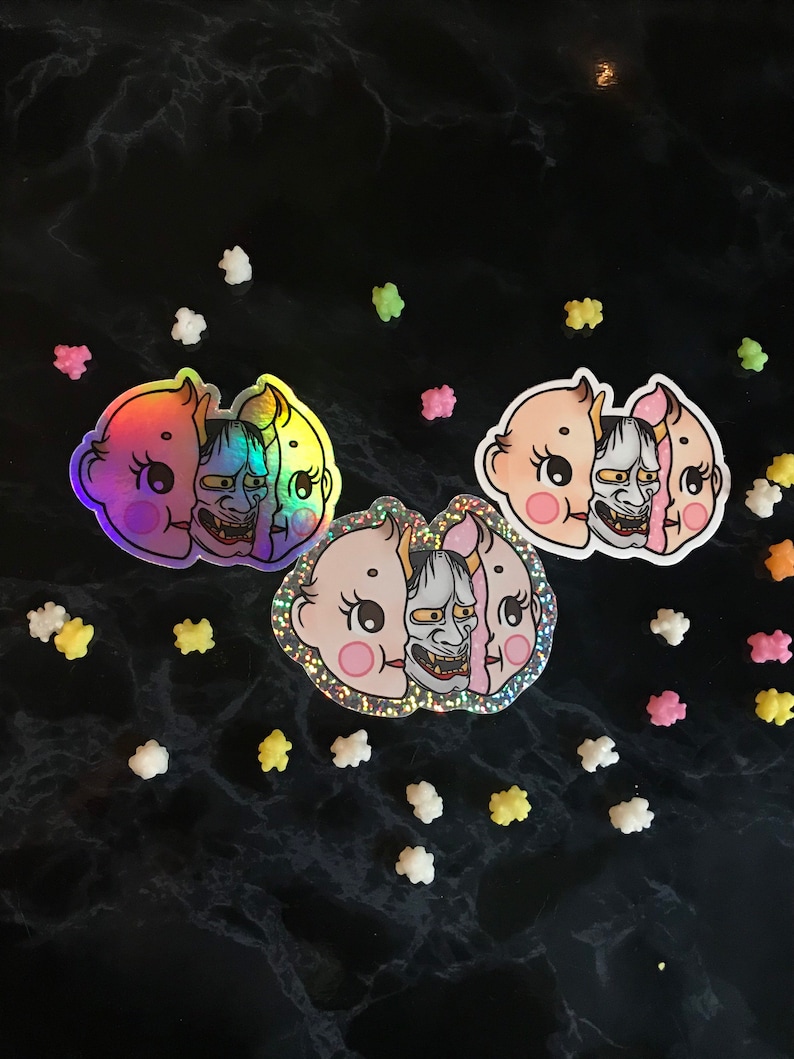 May include: Three iridescent stickers featuring a baby face with a demon mask. The stickers are on a black background with white and pink sprinkles.
