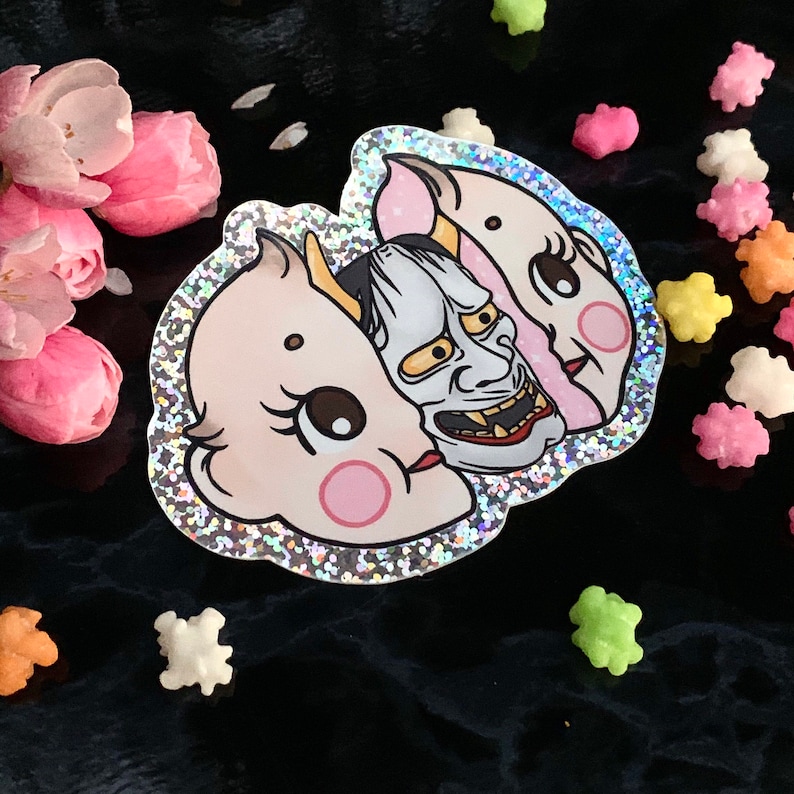 May include: A sticker with a glittery background featuring a split face design. One side is a cute baby doll face with pink cheeks and the other side is a traditional Japanese demon mask with sharp teeth and a fierce expression.