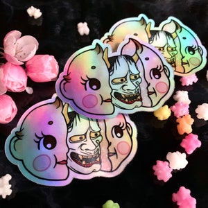 May include: Three iridescent stickers featuring a cute cartoon character with a demon mask. The stickers are holographic and have a rainbow effect.