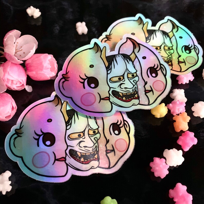 May include: Three iridescent stickers featuring a cute cartoon character with a demon mask. The stickers are holographic and have a rainbow effect.