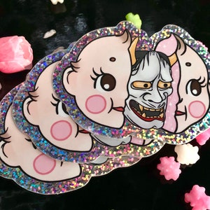May include: A set of four holographic stickers featuring a baby face with a Japanese demon mask. The stickers have a glittery finish.
