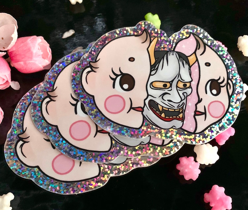 May include: A set of four holographic stickers featuring a baby face with a Japanese demon mask. The stickers have a glittery finish.