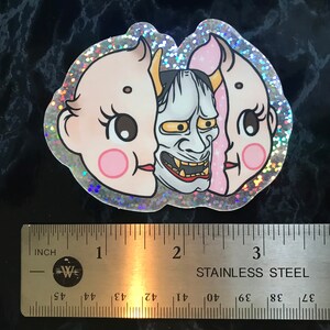 May include: A holographic sticker with a pink and white baby face on one side and a blue and white oni mask on the other side. The sticker is about 2 inches wide.