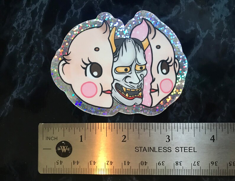 May include: A holographic sticker with a pink and white baby face on one side and a blue and white oni mask on the other side. The sticker is about 2 inches wide.