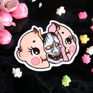 May include: A sticker featuring a split-face design. One side is a cute baby doll with pink cheeks and the other side is a traditional Japanese demon mask with a fierce expression.