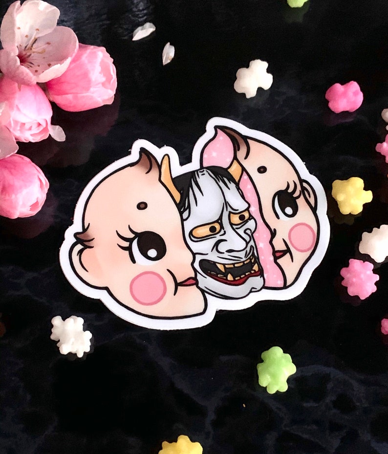 May include: A sticker featuring a split-face design. One side is a cute baby doll with pink cheeks and the other side is a traditional Japanese demon mask with a fierce expression.