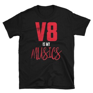 Può includere: T-shirt nera con la scritta rossa "V8 IS MY MUSICS". Il "V8" è in grassetto, con un font a blocchi, mentre "IS MY MUSICS" è in corsivo. La maglietta è a girocollo e maniche corte.