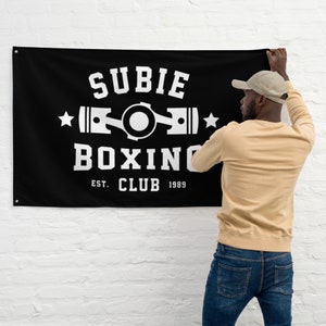 May include: A black flag with a white logo that says "SUBIE BOXING CLUB EST. 1989" with a stylized image of a piston and a circle in the center. A person is hanging the flag on a white brick wall.