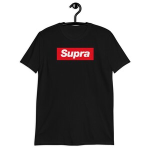 Op de afbeelding: Zwart t-shirt met het woord "Supra" in witte letters in een rode rechthoek. Het t-shirt hangt aan een zwarte hanger.