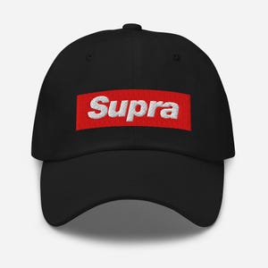 May include: Black baseball cap featuring the word "Supra" in white, embroidered on a red rectangular patch. The cap has a curved brim and a structured crown. A classic, casual accessory.
