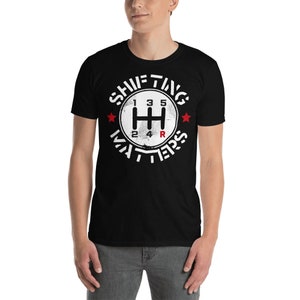 May include: Black t-shirt with a white circle graphic featuring a stylised gear shift pattern and the text "Shifting Matters".