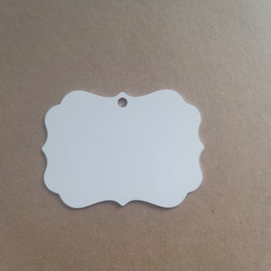May include: White blank ornament with a hole at the top for hanging. The ornament is shaped like a rectangle with rounded corners and a scalloped edge.