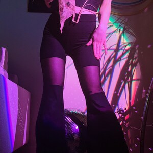 May include: A person wearing a black lace top with a gold chain detail, black trousers with a flared leg, and a sheer pink scarf. The person is standing in front of a disco ball and a wall with a purple and blue light projection.