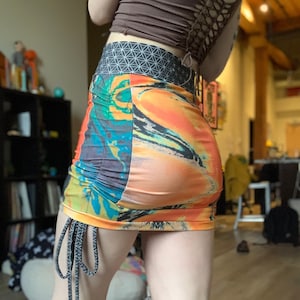 May include: A colorful mini skirt with a tie at the side. The skirt has a geometric pattern waistband and a bright orange, blue, and yellow abstract print.