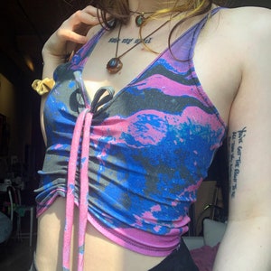 May include: A woman wearing a blue and pink tie-dye crop top with a knotted front. The top has a black and white abstract pattern. The woman has a tattoo on her arm that says "You've got the right vibe" and another tattoo on her chest that says "save my soul".