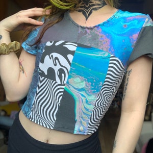 May include: A cropped t-shirt with a patchwork design featuring abstract patterns in black, white, blue, and pink. The shirt has a crew neck and short sleeves.
