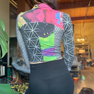 Puede incluir: Top corto de manga larga con cuello alto. El top presenta un diseño de patchwork con patrones geométricos y florales en negro, blanco, rosa, morado y verde. Las mangas son grises con un patrón geométrico blanco. Combinado con leggings negros.