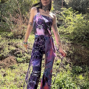 以下が含まれることがあります： 紫とピンクのタイダイ柄のワンピースを着た人。ジャンプスーツは黒いチェーン柄と胸元に白いストラップが付いています。白い帽子をかぶり、腕にタトゥーを入れています。