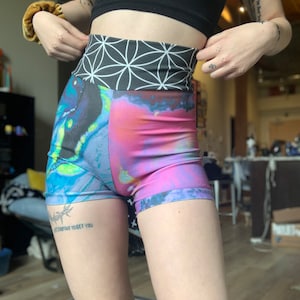 Pode incluir: Shorts de cintura alta, coloridos e estampados com uma cintura preta que apresenta um padrão geométrico. Os shorts têm um esquema de cores azul, rosa e roxo com uma estampa semelhante a uma galáxia.