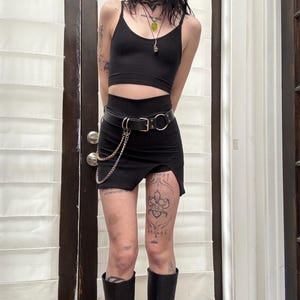 Pode incluir: Uma pessoa vestindo um top preto curto, uma minissaia preta com um cinto de corrente prateada e botas pretas de cano alto. A pessoa tem tatuagens nas pernas.