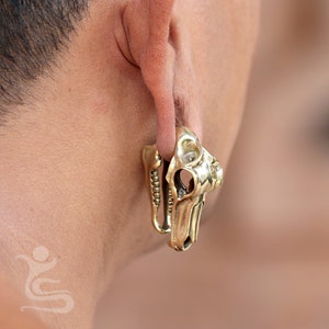 May include: A gold-coloured skull-shaped earring with intricate details. The earring is worn in the earlobe of a person.