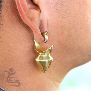 May include: A gold-colored ear gauge, featuring a curved top and a diamond-shaped bottom. The jewelry is worn in a stretched earlobe, showcasing its unique design and metallic sheen. The gauge is a bold accessory.