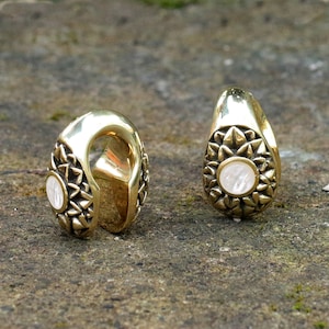 May include: Two gold-toned ear gauges with a white, iridescent stone set in the center of each. The gauges have a detailed, ornate design.