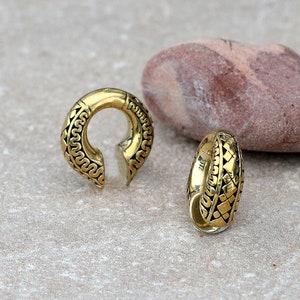 May include: Two gold-colored metal ear gauges with intricate geometric designs. The gauges are circular and have a slightly curved shape.