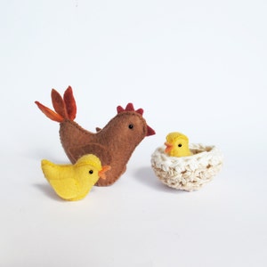 May include: A felt hen, two yellow chicks, and a crocheted nest. The hen is brown with a red comb and orange tail feathers. One chick stands beside the hen, and the other sits in the nest. The nest is cream-colored.