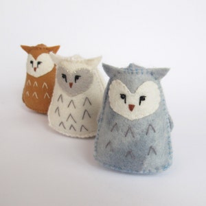 May include: Three felt owl ornaments in brown, white, and blue. Each owl has a simple embroidered face and a pattern of small triangles on its body.
