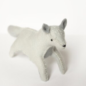 May include: A gray felt toy wolf with stitched details, standing on a white surface.