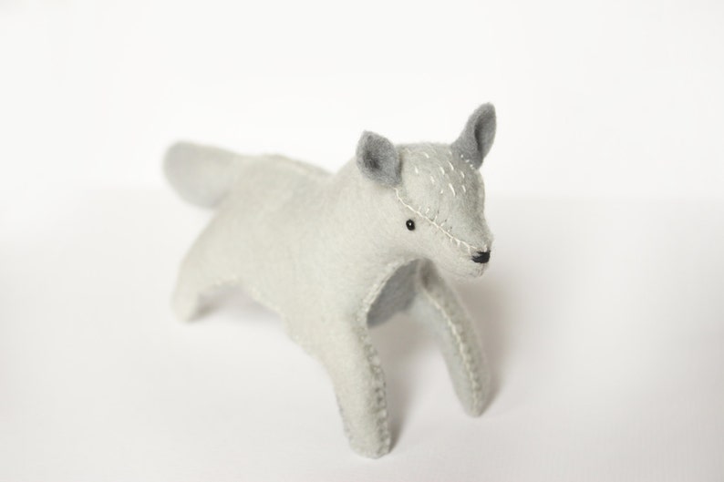 May include: A gray felt toy wolf with stitched details, standing on a white surface.