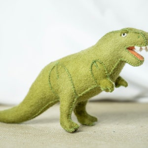 May include: Green felt T-Rex dinosaur toy with stitched details. The dinosaur has a wide open mouth with white teeth and an orange tongue.